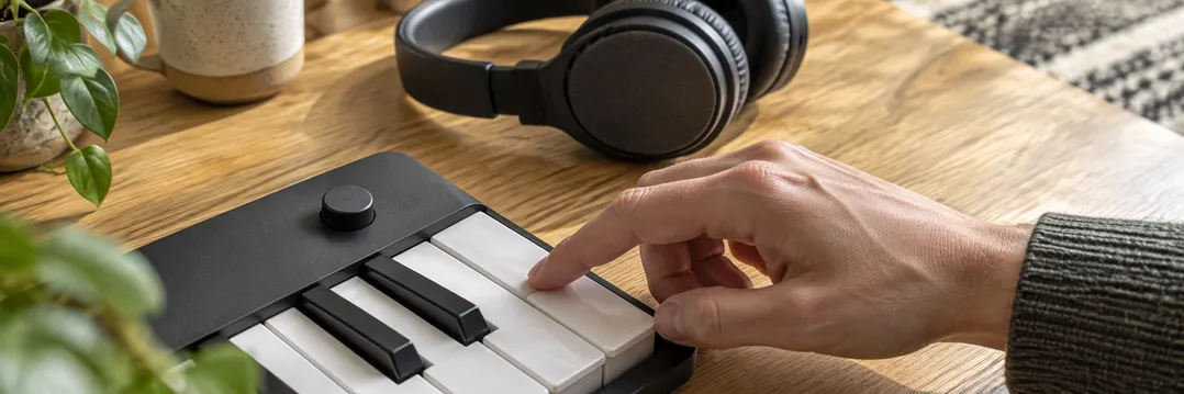 Eine Hand probiert eine Keyboardtaste aus; dahinter liegen Kopfhörer auf einem Holztisch.